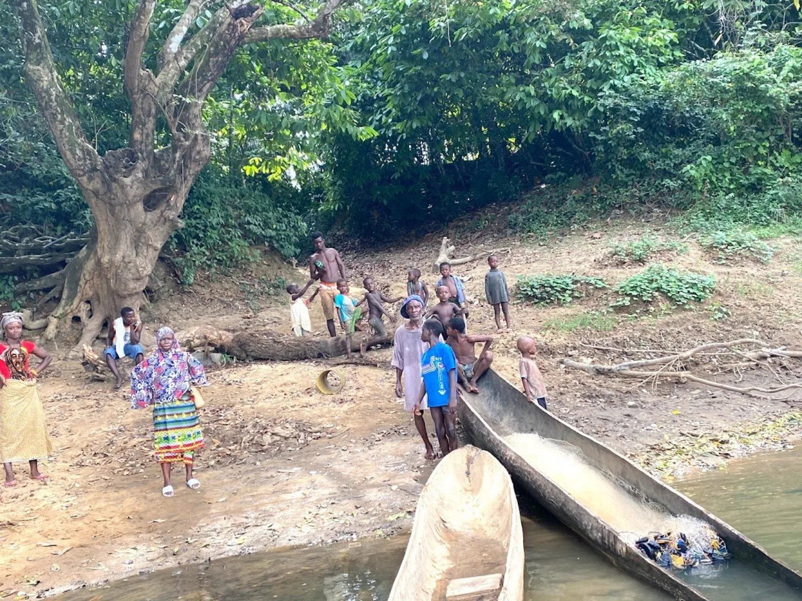 Families beside wooden canoes on a riverbank in a Gbema partner village